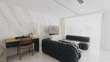 Una habitación de hotel moderna con pared de mármol, un escritorio de madera, un sofá negro con capitoné, un televisor de pantalla plana y un ventanal con cortinas traslúcidas.