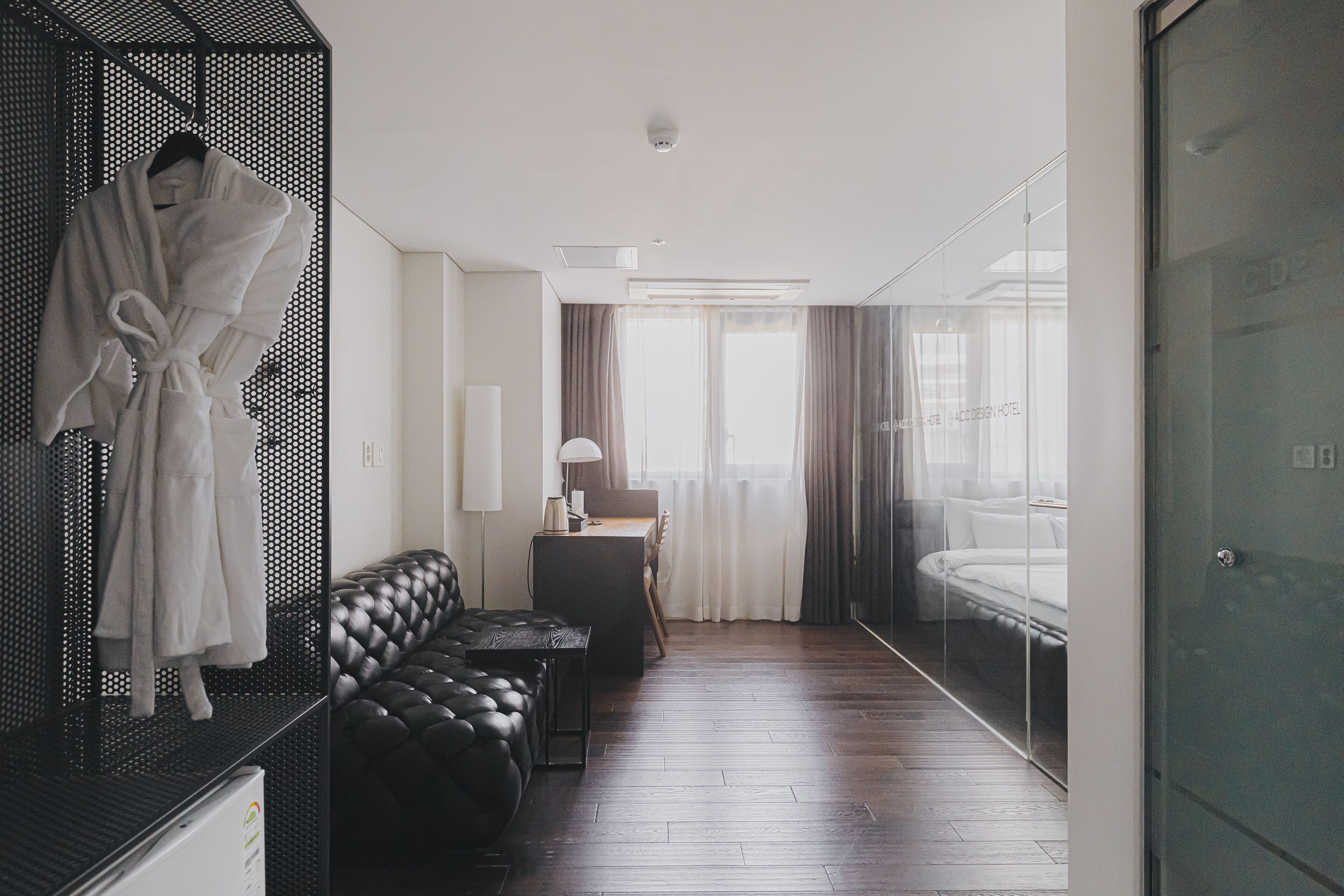 Una habitación de hotel moderna con sofá de cuero, mesita de noche, cama con cabecera de vidrio y bata colgada en un perchero.