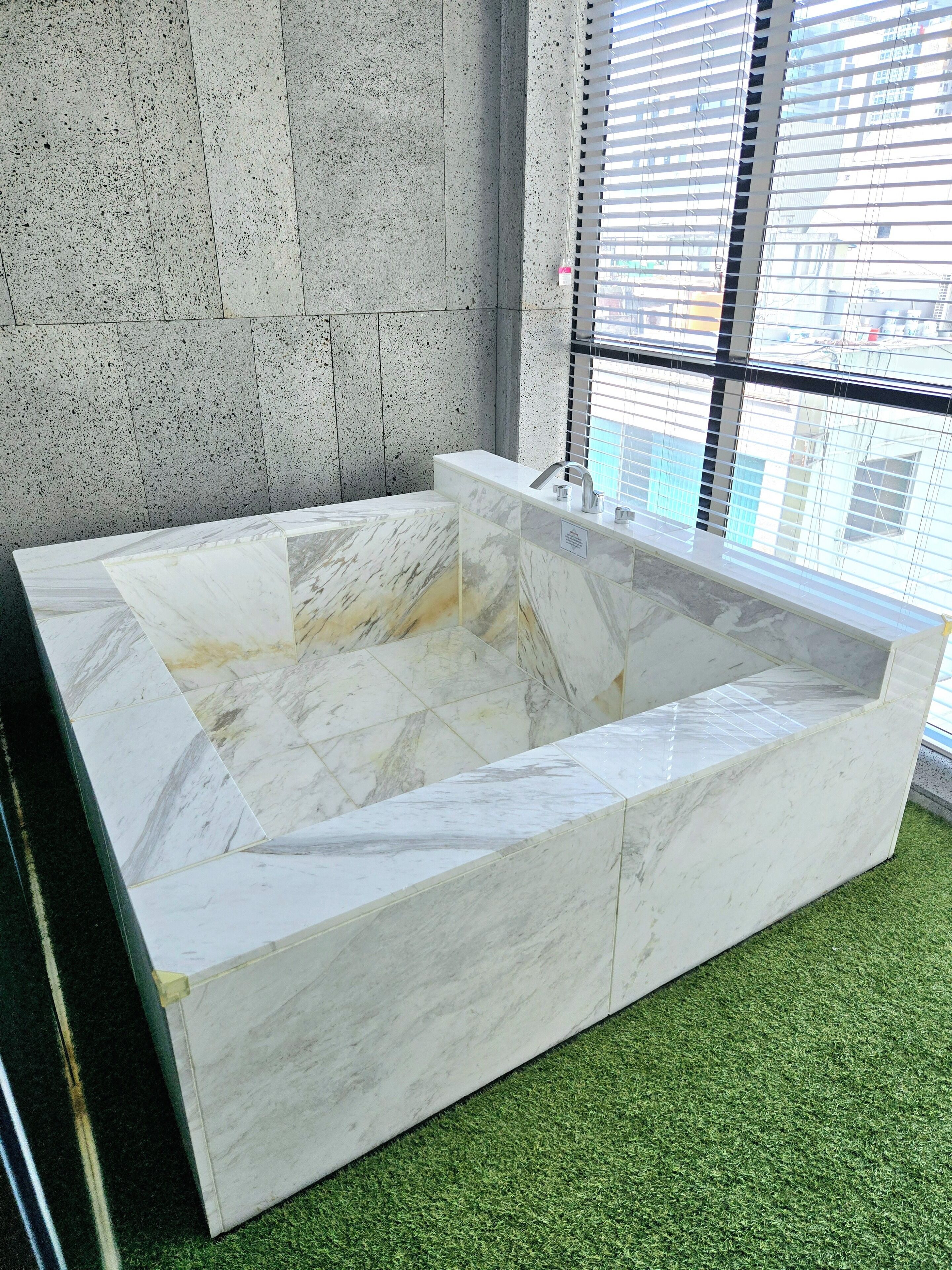 A rectangular marble bathtub with a built-in faucet, set against a concrete wall and a window with blinds.
