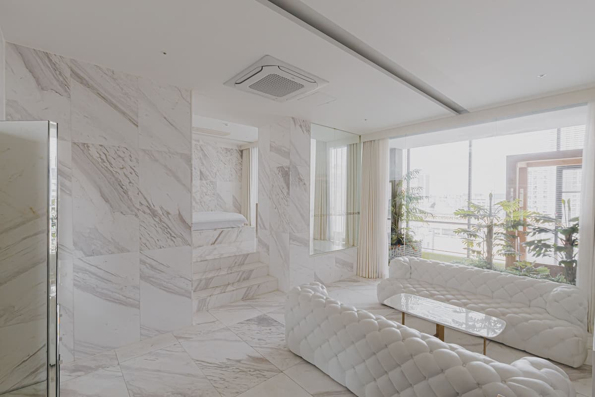 Una habitación moderna con suelo de mármol, cabina de ducha de vidrio, zona de estar con cojines blancos y un amplio ventanal con vistas a la ciudad.