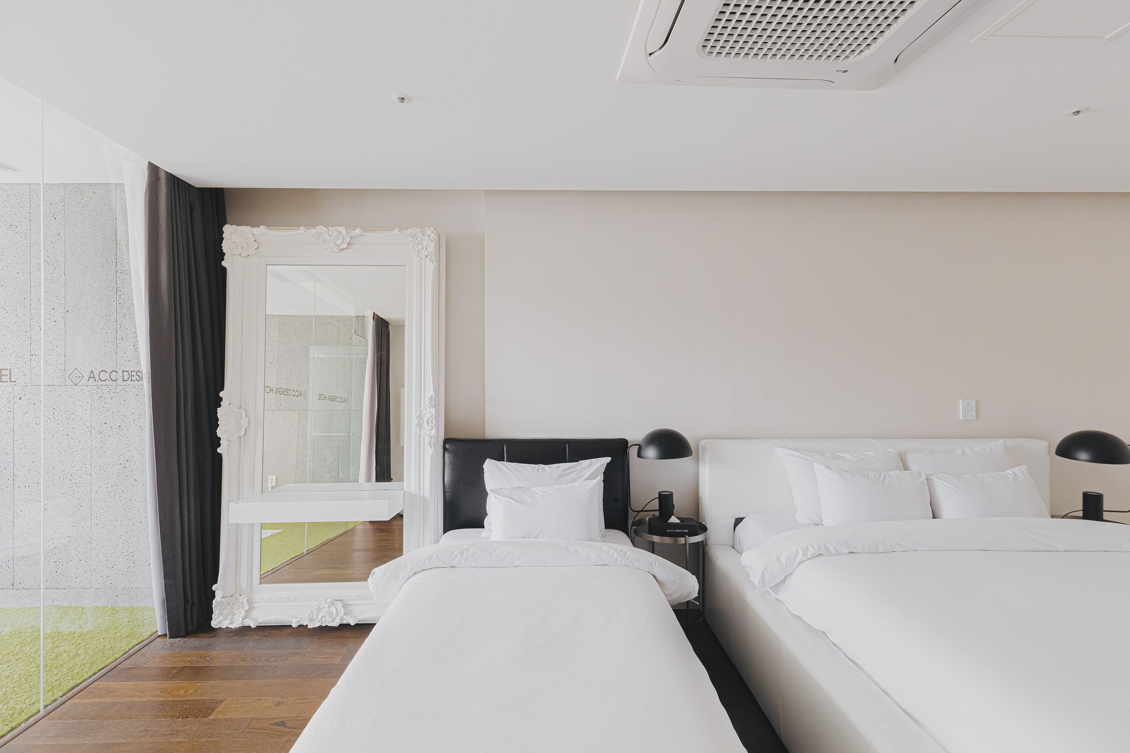 Habitación de hotel con dos camas, un espejo grande, una mesita auxiliar y vistas a un espacio exterior arbolado.