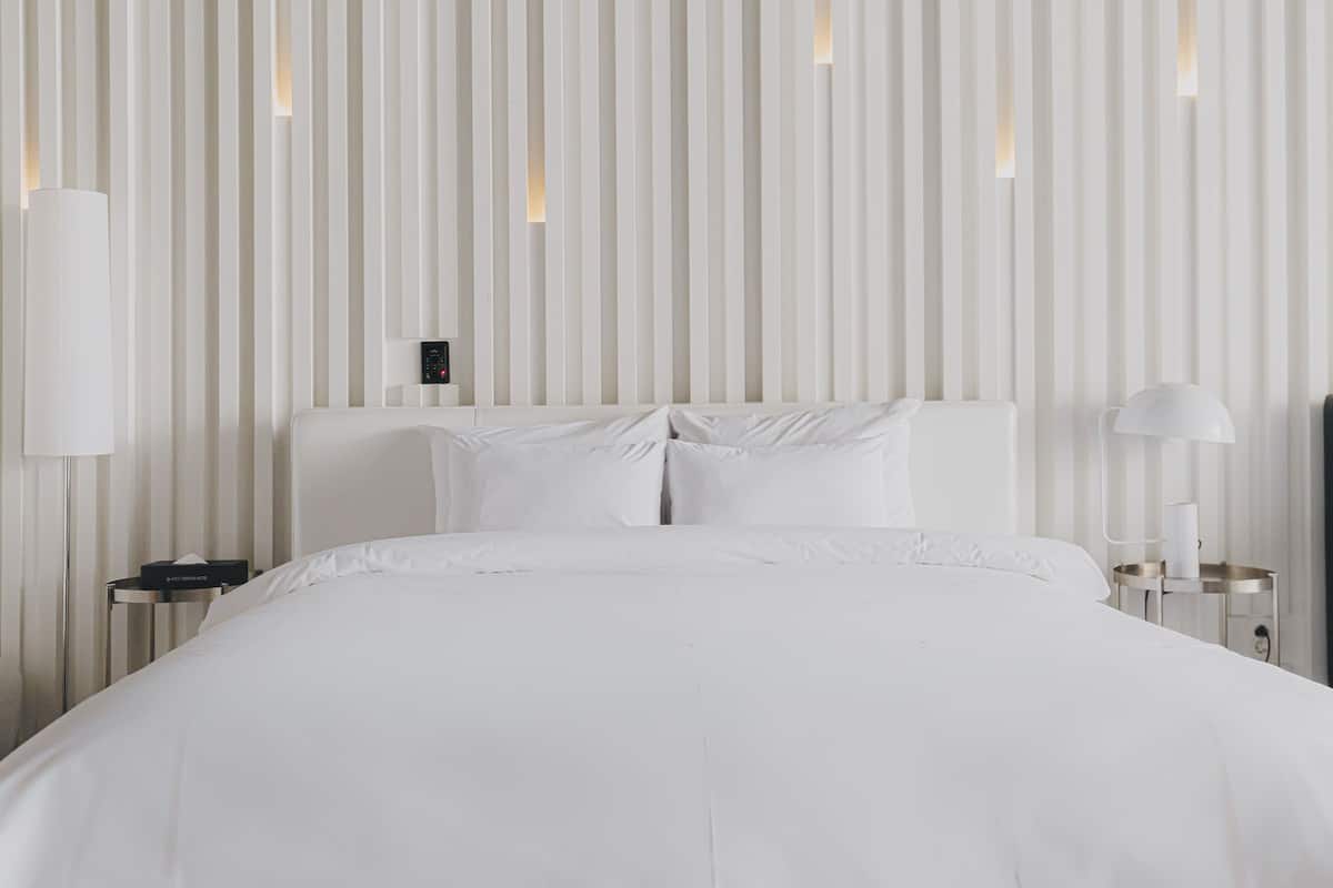Habitación de hotel moderna con una cama grande, ropa de cama blanca, una mesita de noche con lámpara y un panel de control montado en la pared.