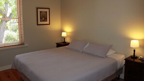 Een net opgemaakt bed met een witte deken en kussens, twee nachtkastjes met lampen, een ingelijste foto aan de muur, en een raam met jaloezieën.