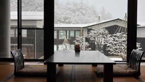 A modern dining area with a large table and two chairs, a window with a view of snow-covered trees and a building. - Hotel Kikori (Hida)