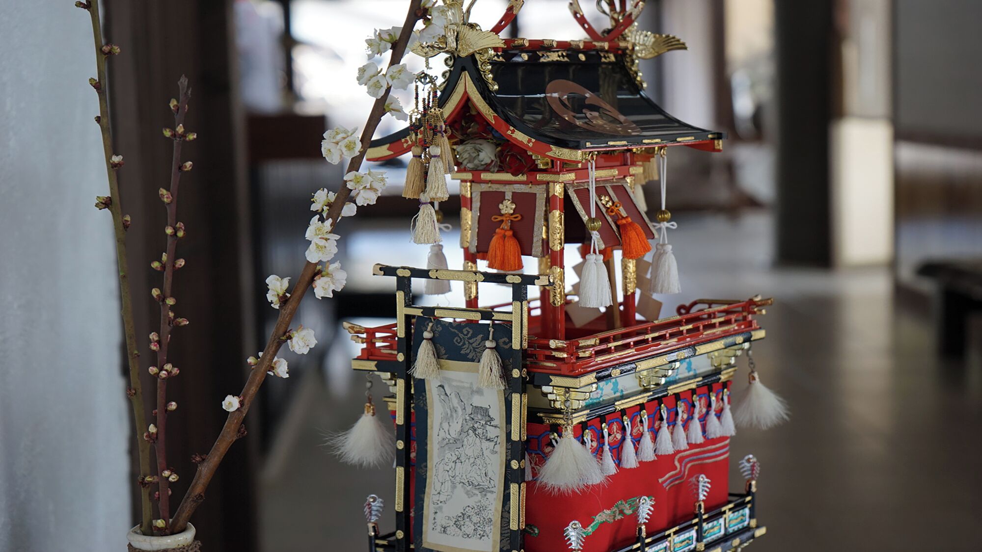 Un bâtiment japonais traditionnel, avec une façade rouge et dorée, des décorations florales blanches et un toit noir.