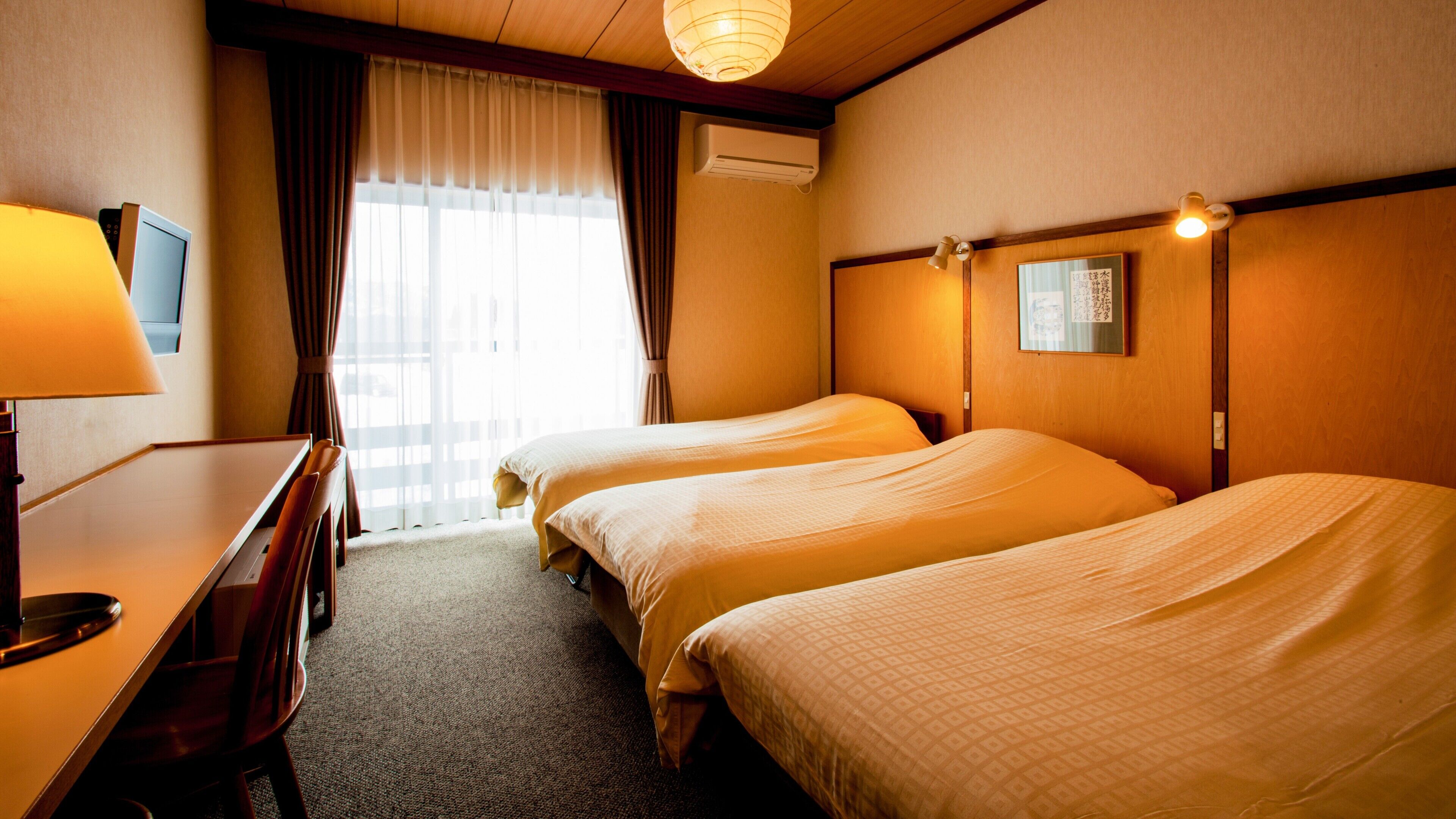 A hotel room with two beds, a desk, a chair, an air conditioning unit, and a window with curtains.