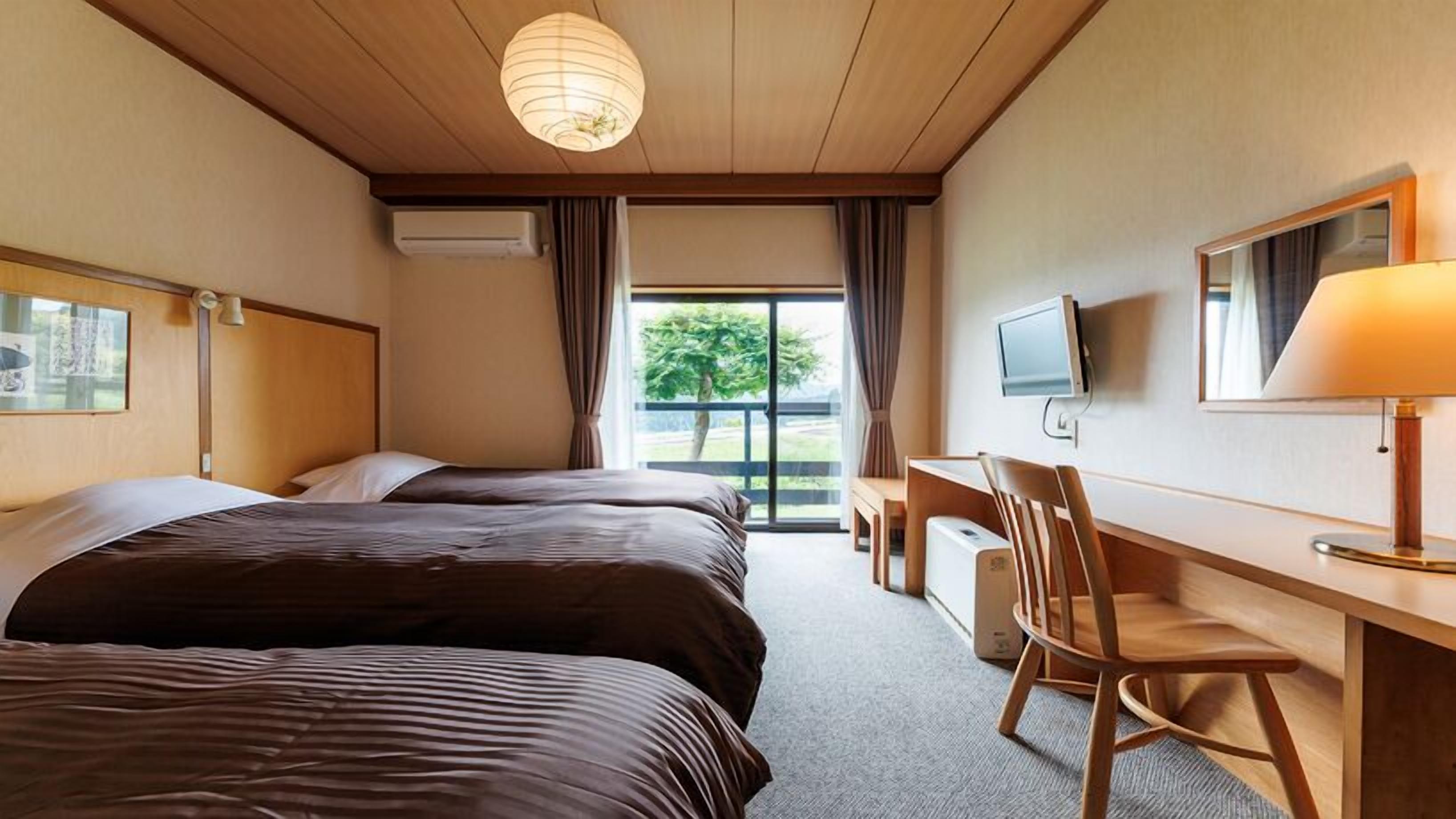 Habitación de hotel con tres camas, un escritorio con silla, televisión y un ventanal con cortinas.
