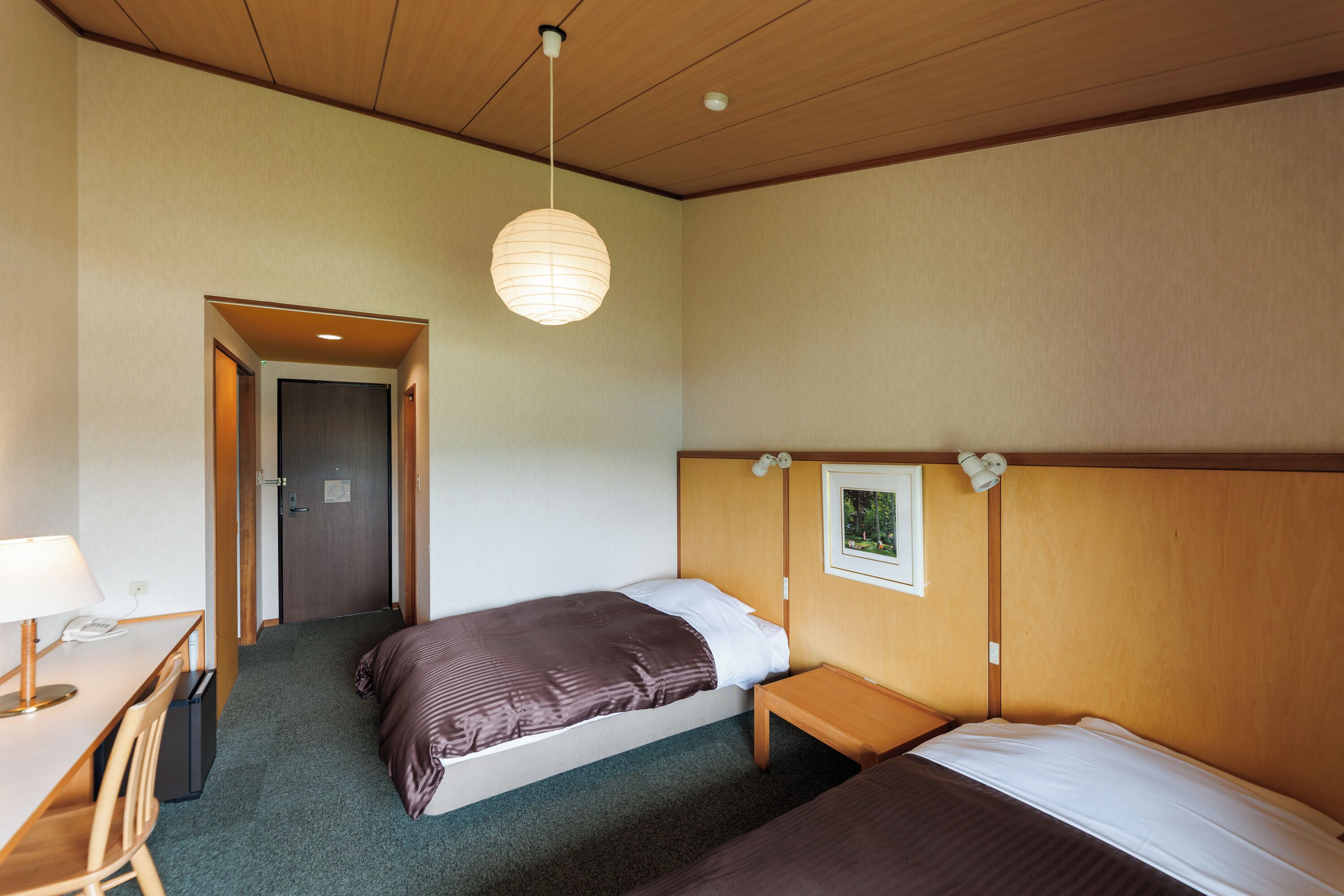 A hotel room with two beds, a desk with a lamp, and a wooden ceiling.