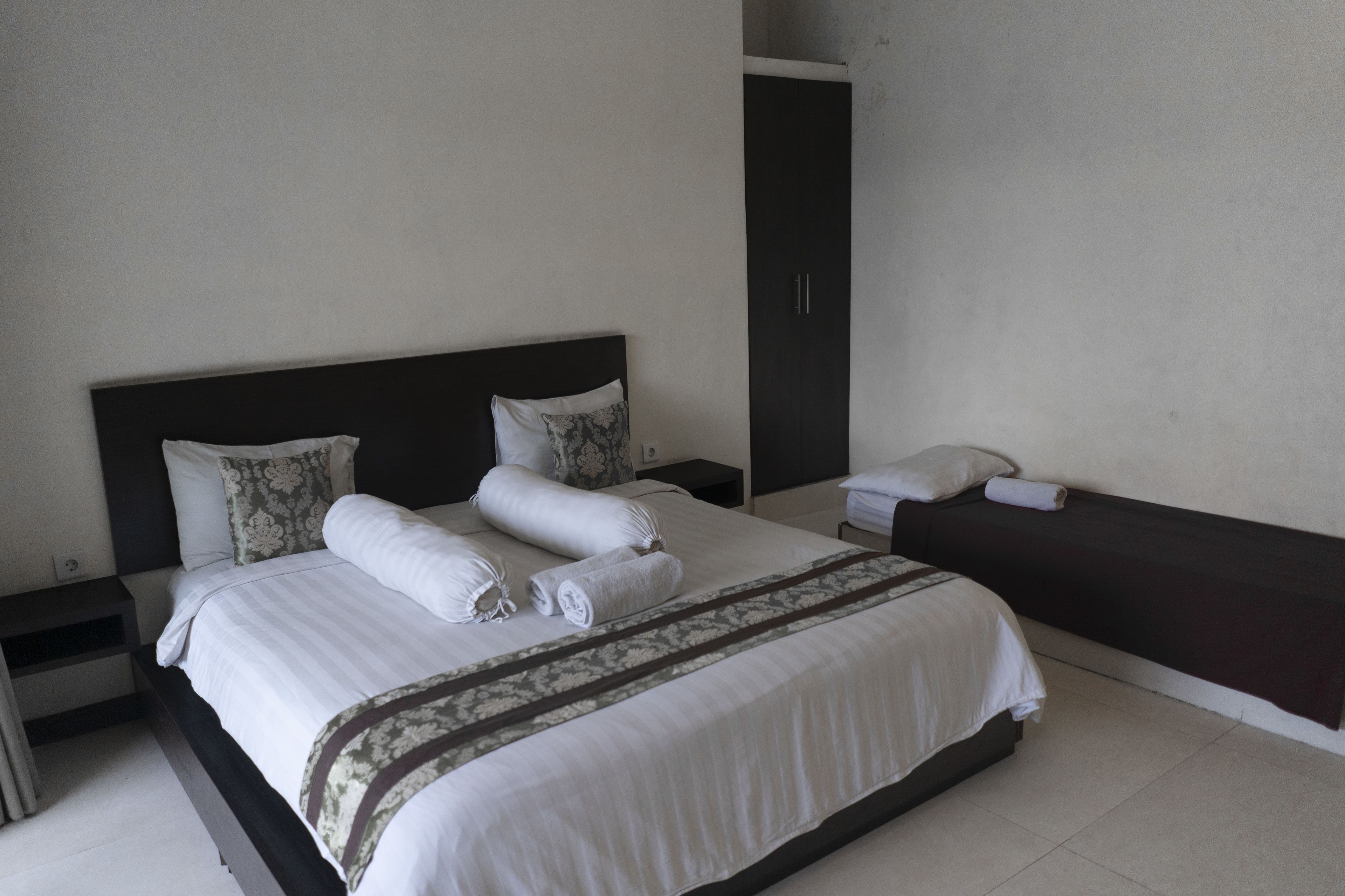 A neatly made bed with white linens and a decorative throw, a bedside table with a lamp, and a dark-colored bench against the wall.