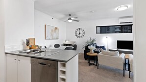 A modern kitchen with a dishwasher, a dining area with a table and chairs, a sofa, a TV mounted on the wall, and a ceiling fan.