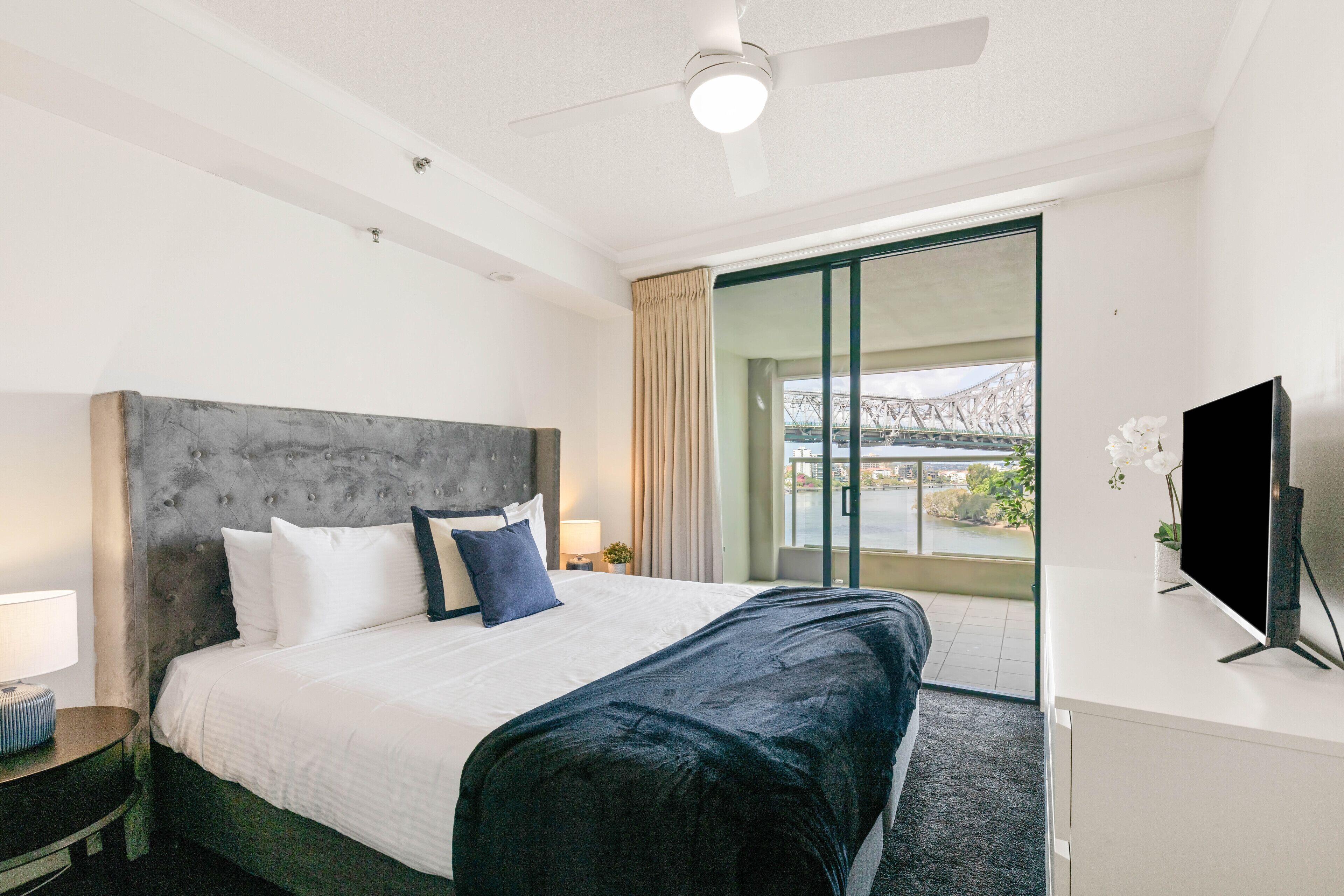 A hotel room with a large bed, a flat-screen TV, a desk, and a balcony with a view of a bridge.