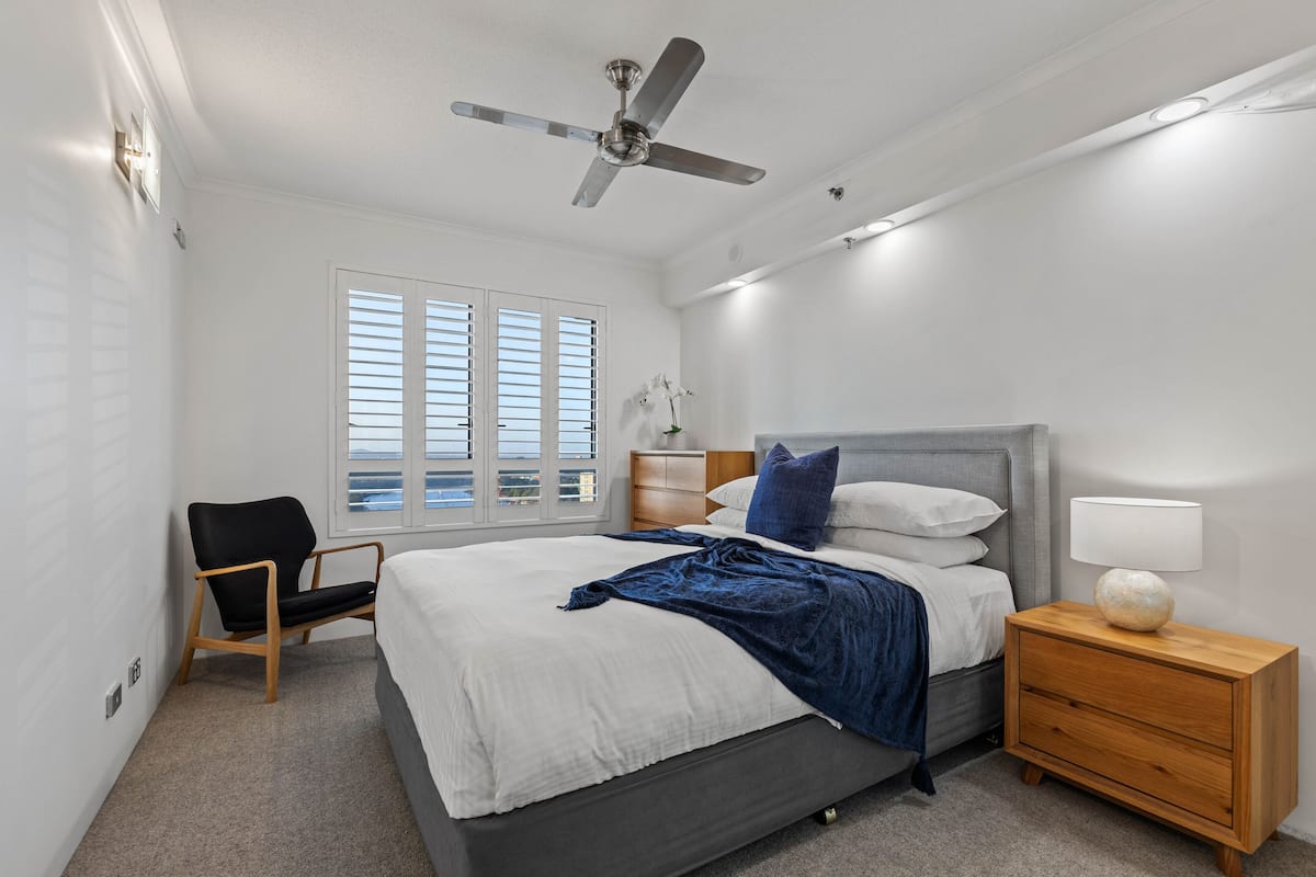 A bedroom with a bed, a chair, a nightstand, a ceiling fan, and a window with blinds.
