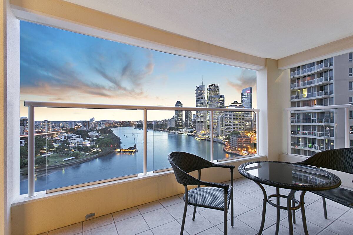 A balcony with a view of a river and cityscape, featuring a round table and two chairs.