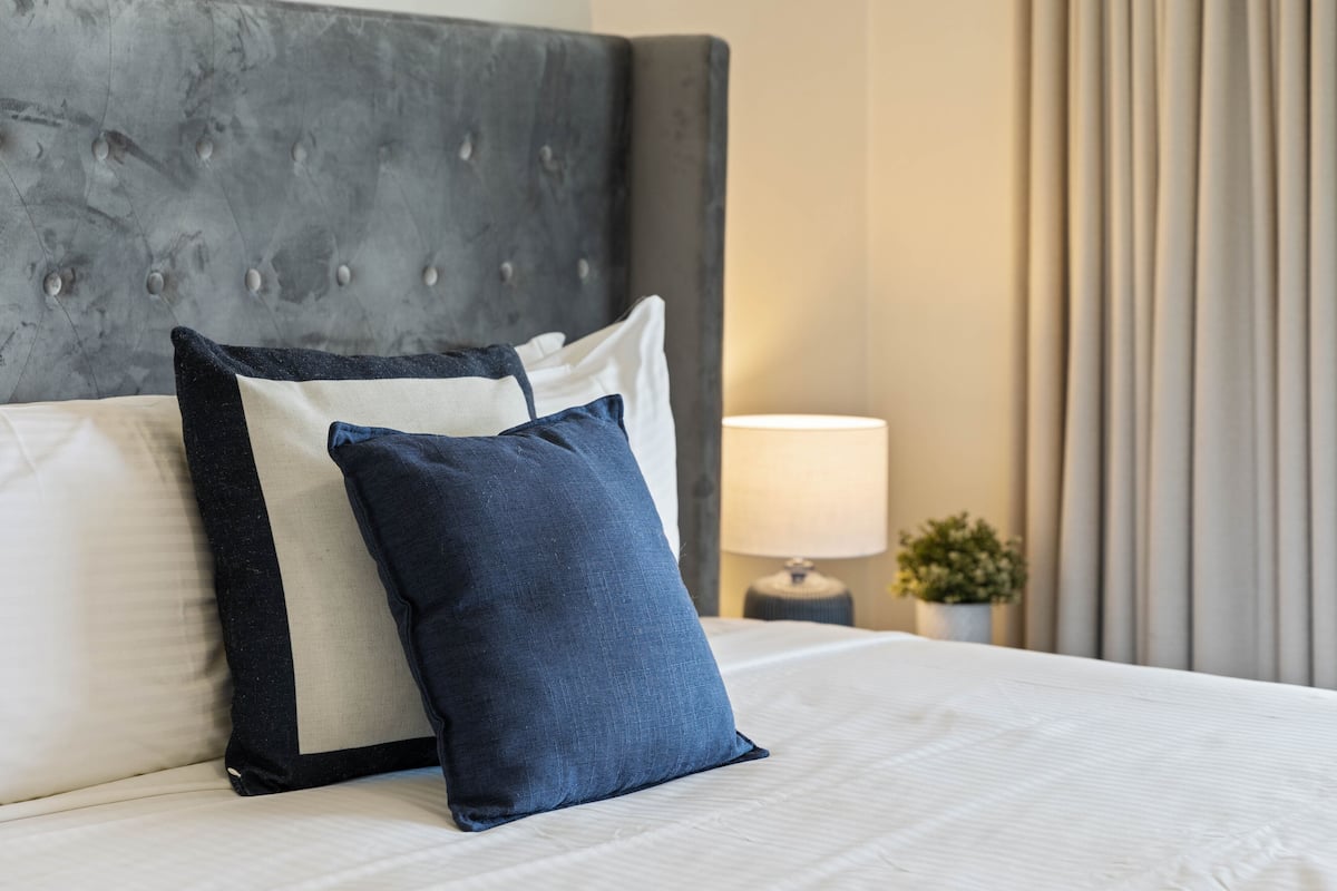 A neatly made bed with a grey upholstered headboard, two pillows, a bedside lamp, and a potted plant.