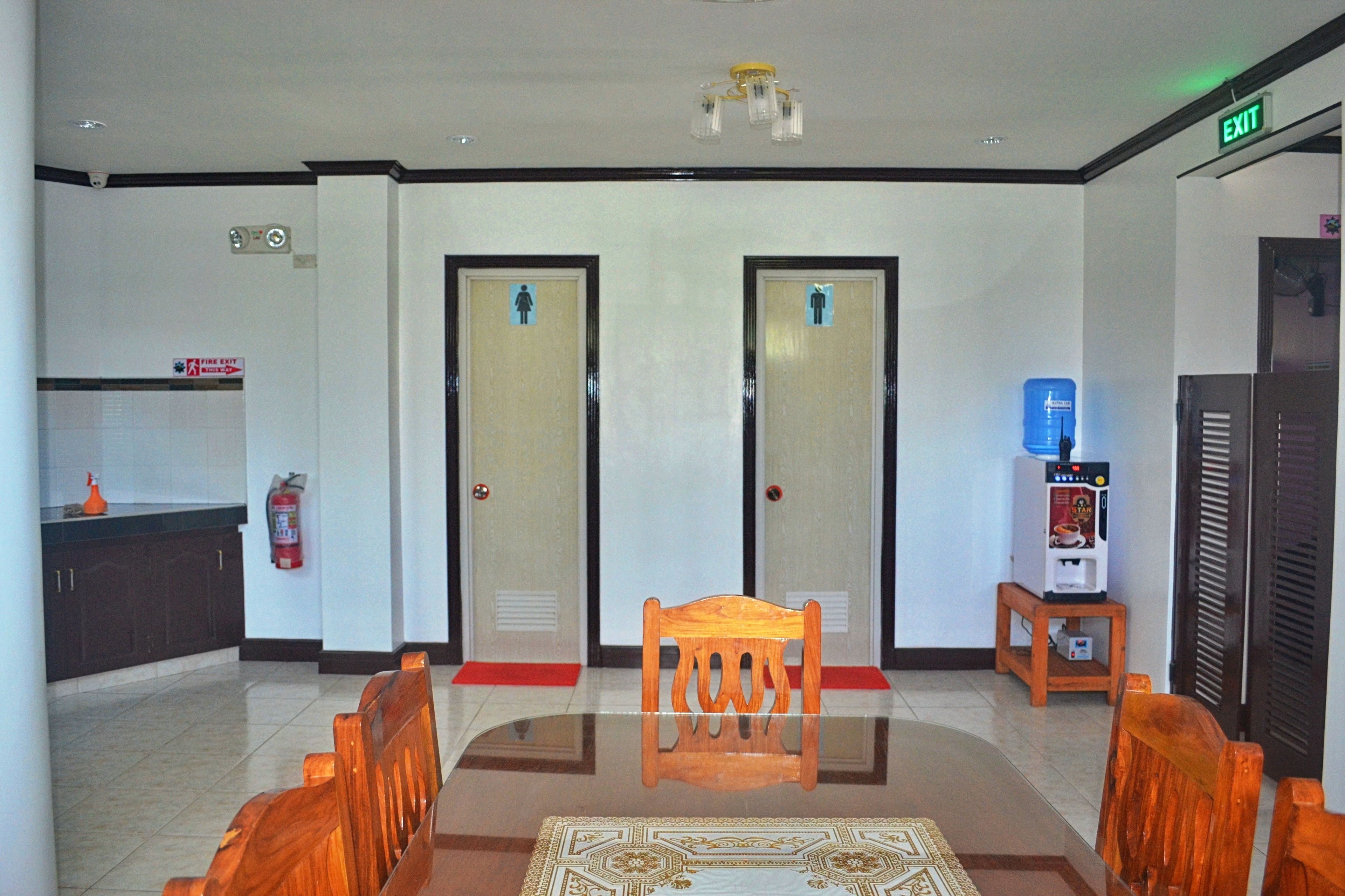 A lobby with a reception desk, two doors with gender symbols, a fire extinguisher, a water dispenser, and a wooden chair.