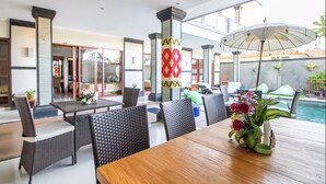 A dining area with a wooden table and wicker chairs, a decorative column, and a pool in the background. - Lebak Bali Nelayan (Canggu)