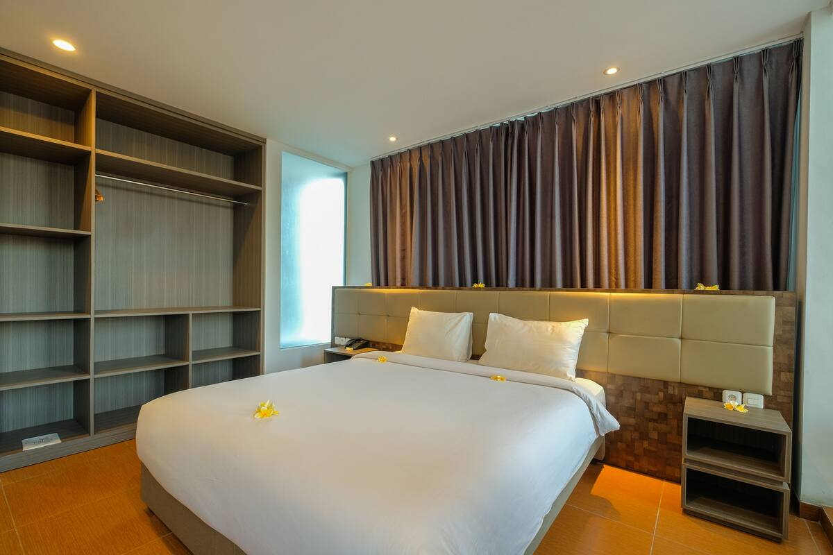 A hotel room with a large bed, a wardrobe, a nightstand, and a headboard with built-in lighting.