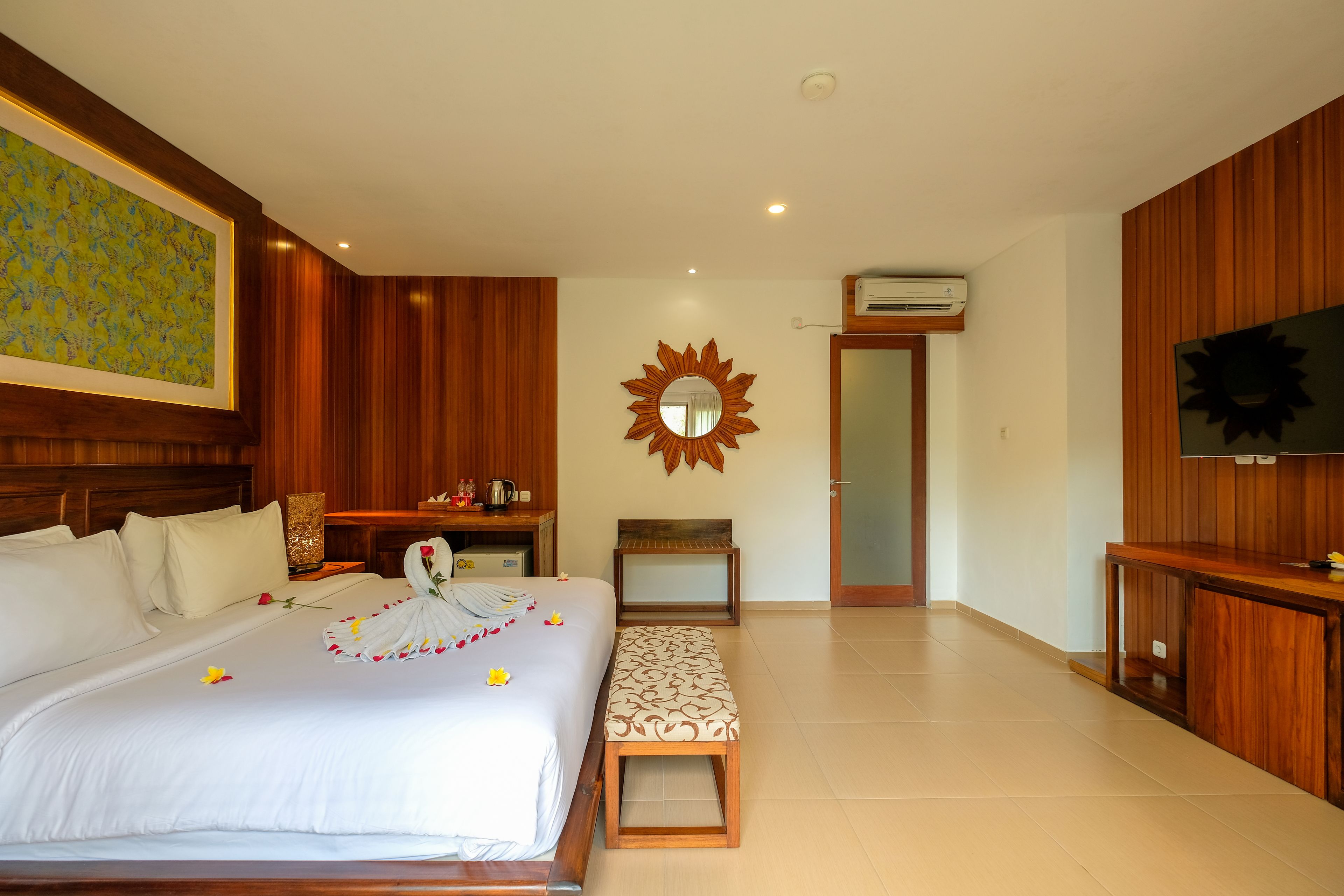 A hotel room with a large bed, a wooden bench, a sun-shaped mirror, a television, and a wooden panel wall.