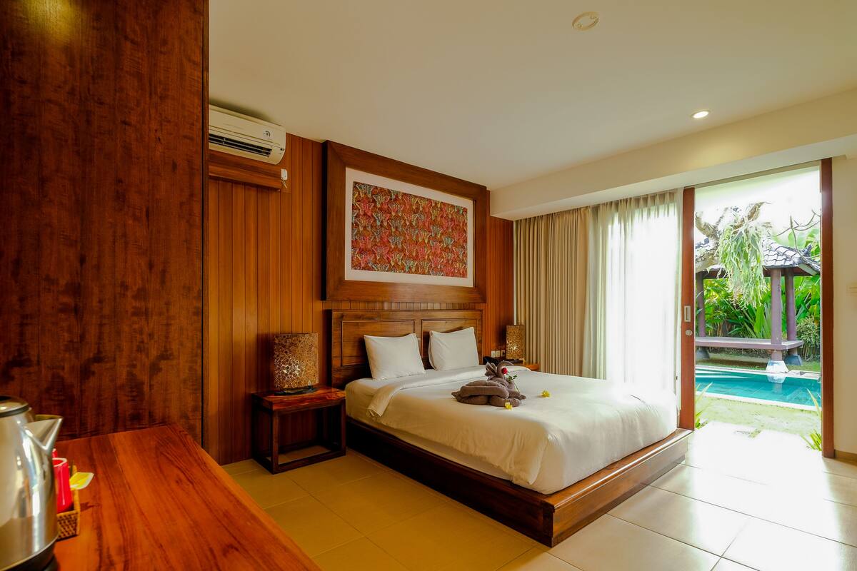 A hotel room with a wooden headboard, a large bed with white linens, a small table with a kettle, and a view of a swimming pool through a glass door.