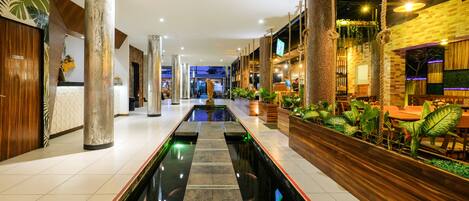 A hotel lobby with a central water feature, wooden columns, and a seating area with plants.