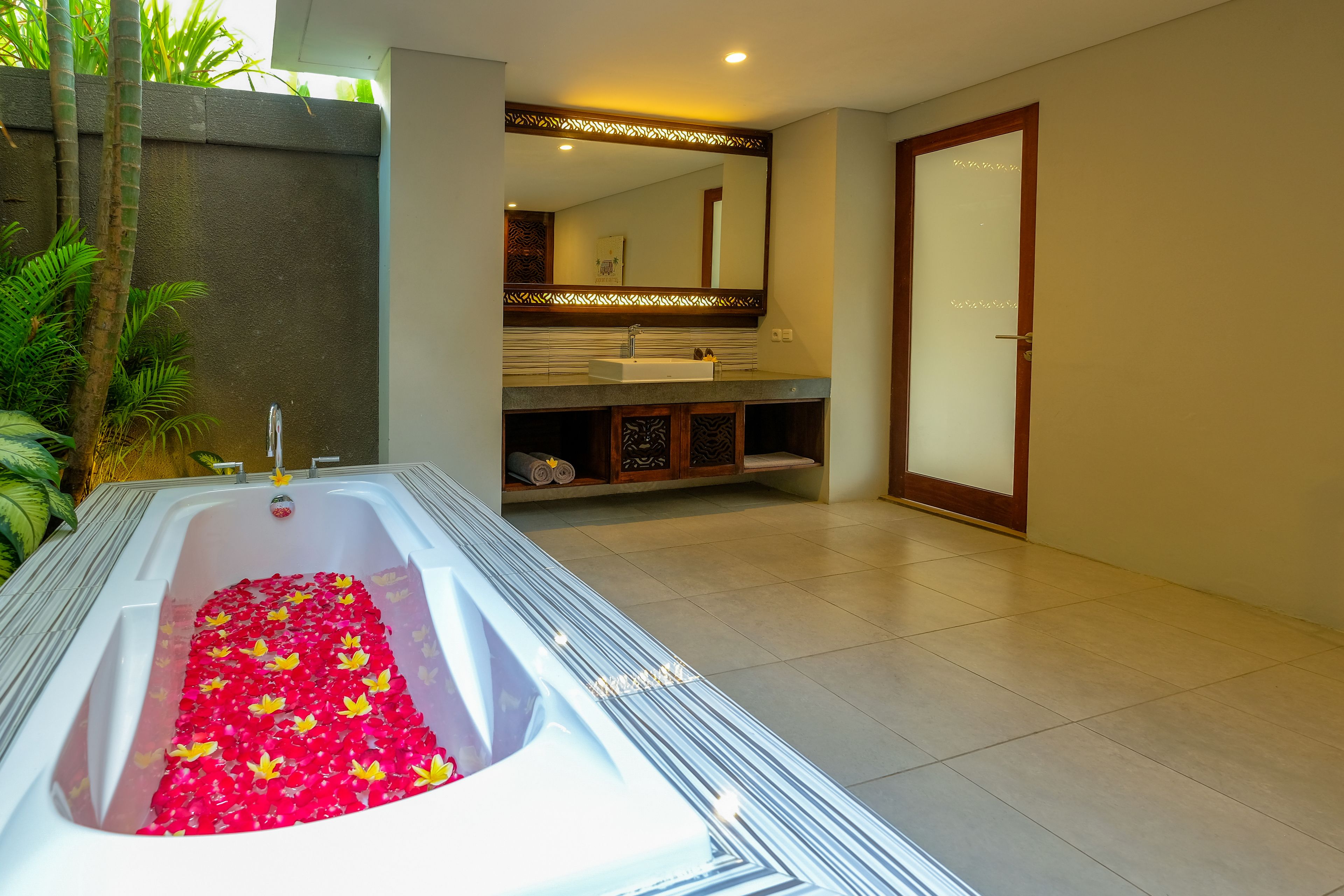 Une salle de bain moderne avec une grande baignoire remplie de pétales de rose, un meuble-lavabo avec un miroir et une porte dotée de panneaux en verre dépoli.