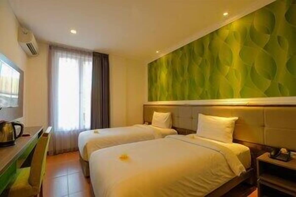 A hotel room with two beds, a green textured wall, a window with curtains, and a bedside table with a lamp.