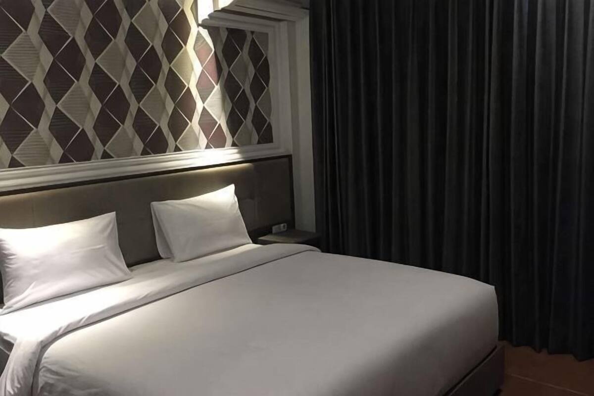 A neatly made bed with white linens, a headboard with a diamond pattern, and a window with curtains.