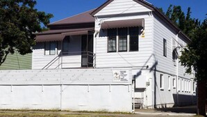 A white two-story building with a brown roof, a covered front porch, and a small front yard. - Gonow Family Backpackers Hostel (West End)