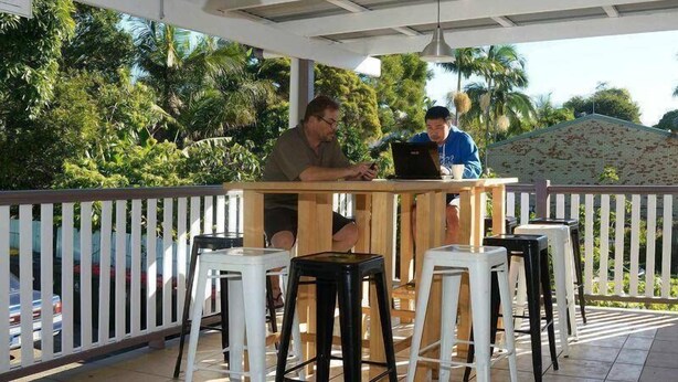 Two people working at a wooden table with a laptop, surrounded by white and black stools, under a covered area with a white railing and palm trees in the background. - Gonow Family Backpackers Hostel (West End)