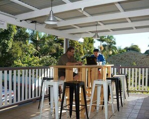 Two people working at a wooden table with a laptop, surrounded by white and black stools, under a covered area with a white railing and palm trees in the background. - Gonow Family Backpackers Hostel (West End)