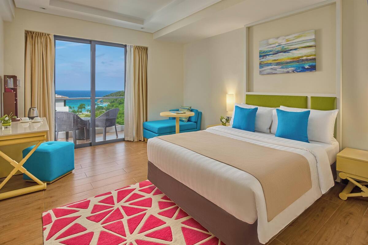 A hotel room with a large bed, a desk with a chair, a small table, and a view of the ocean.