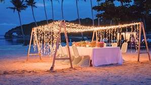 A beachside dining setup with string lights, white tablecloths, and wicker chairs.