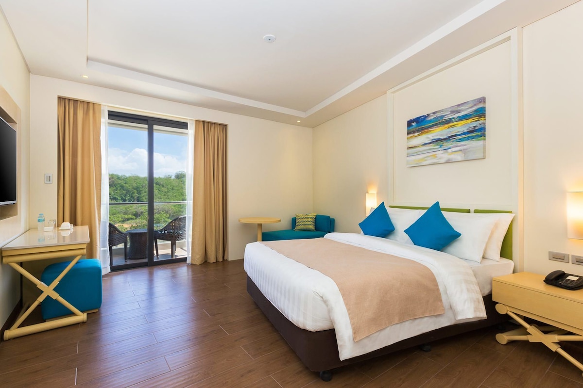 A hotel room with a bed, a desk, a chair, a TV, a balcony, and a painting on the wall.