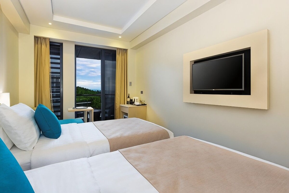 A hotel room with two beds, a TV mounted on the wall, a balcony with curtains, and a small table with a lamp.