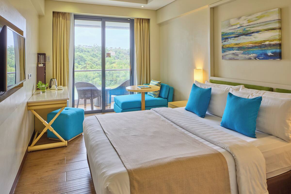 A hotel room with a large bed, a desk with a chair, a small table, and a balcony with a view of greenery.