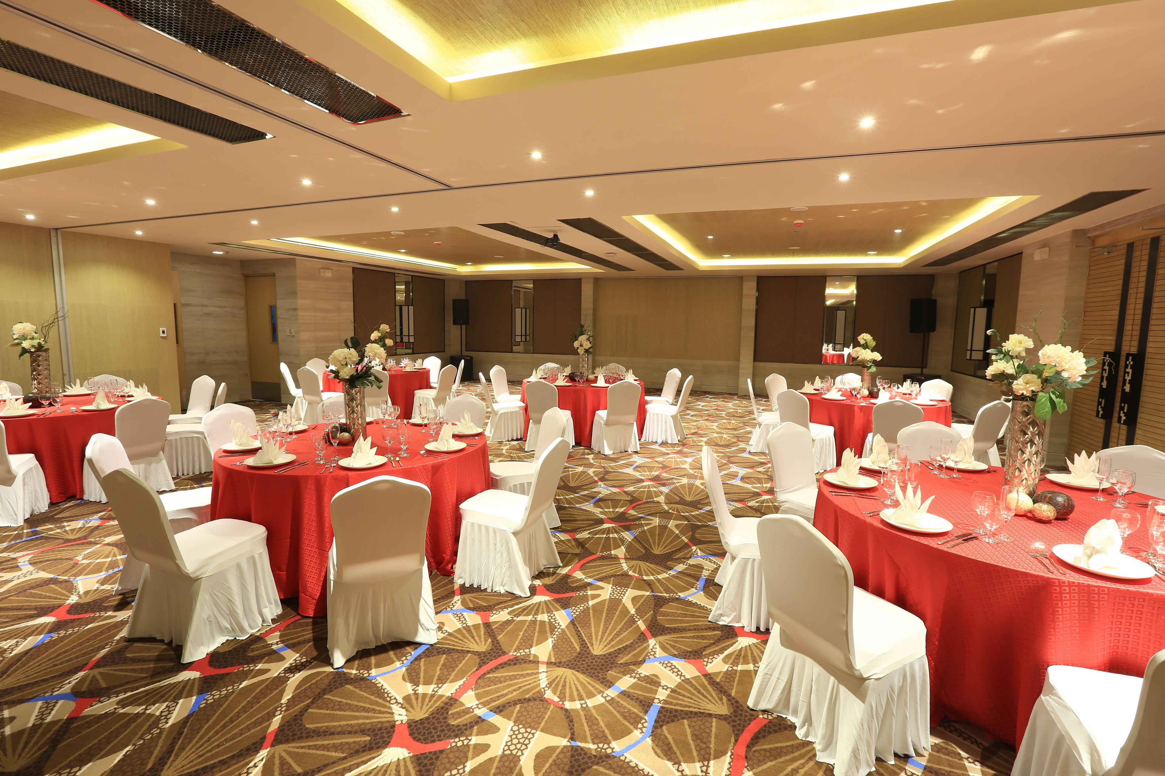 Un salón de banquetes con mesas redondas cubiertas con manteles rojos, sillas blancas y centros de mesa florales.