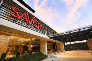 Un hotel moderno con un llamativo letrero de 'SAVOY HOTEL', grandes ventanales y un área de entrada techada.