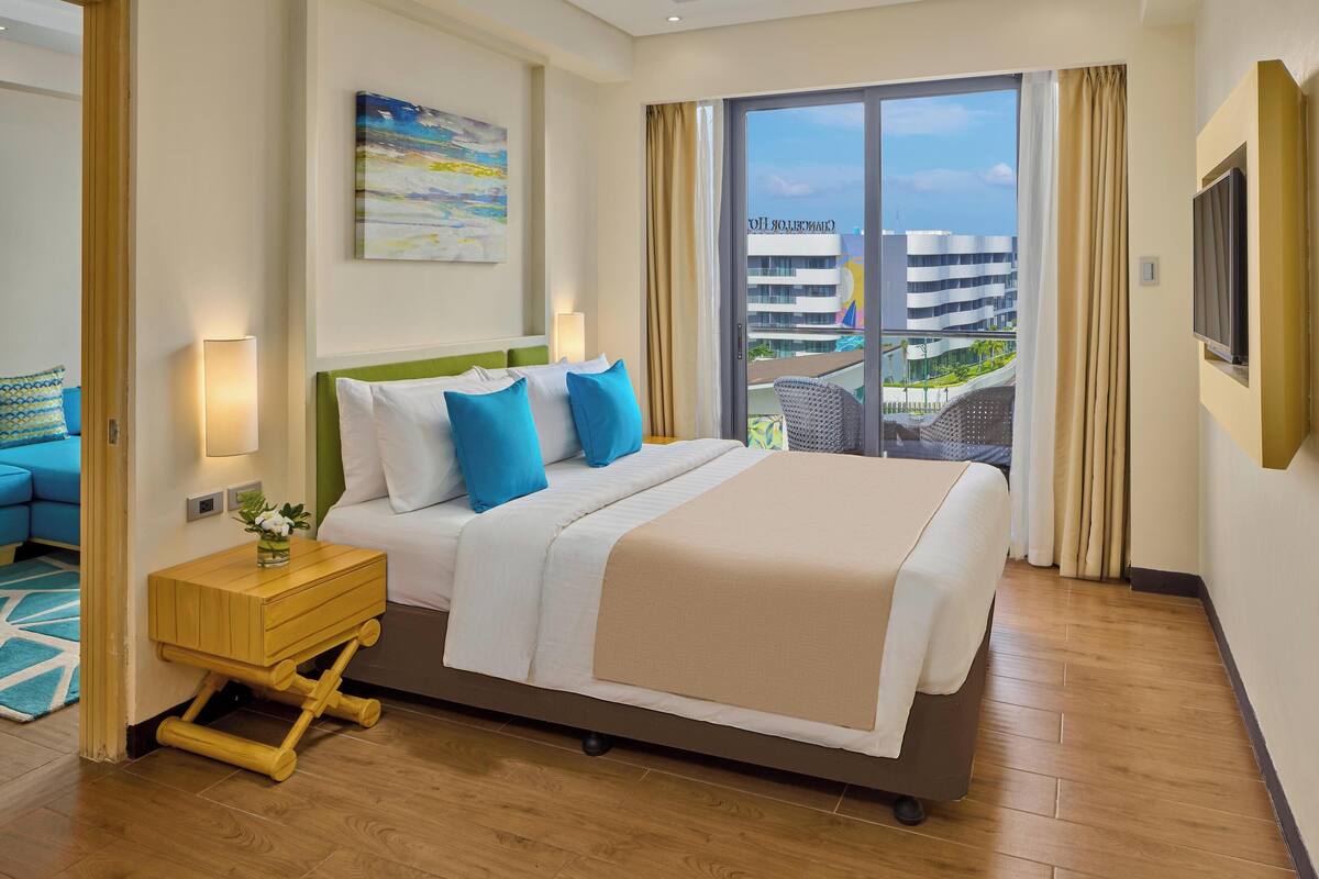 A hotel room with a large bed, a nightstand, a TV, and a view of a building through the window.