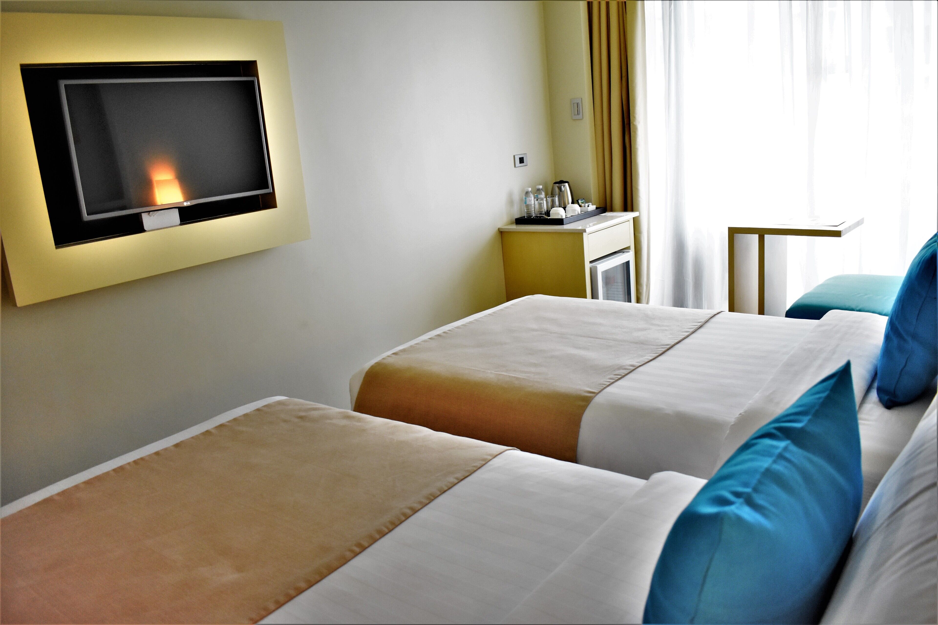 A hotel room with two beds, a TV mounted on the wall, a small table with a kettle, and a window with curtains.