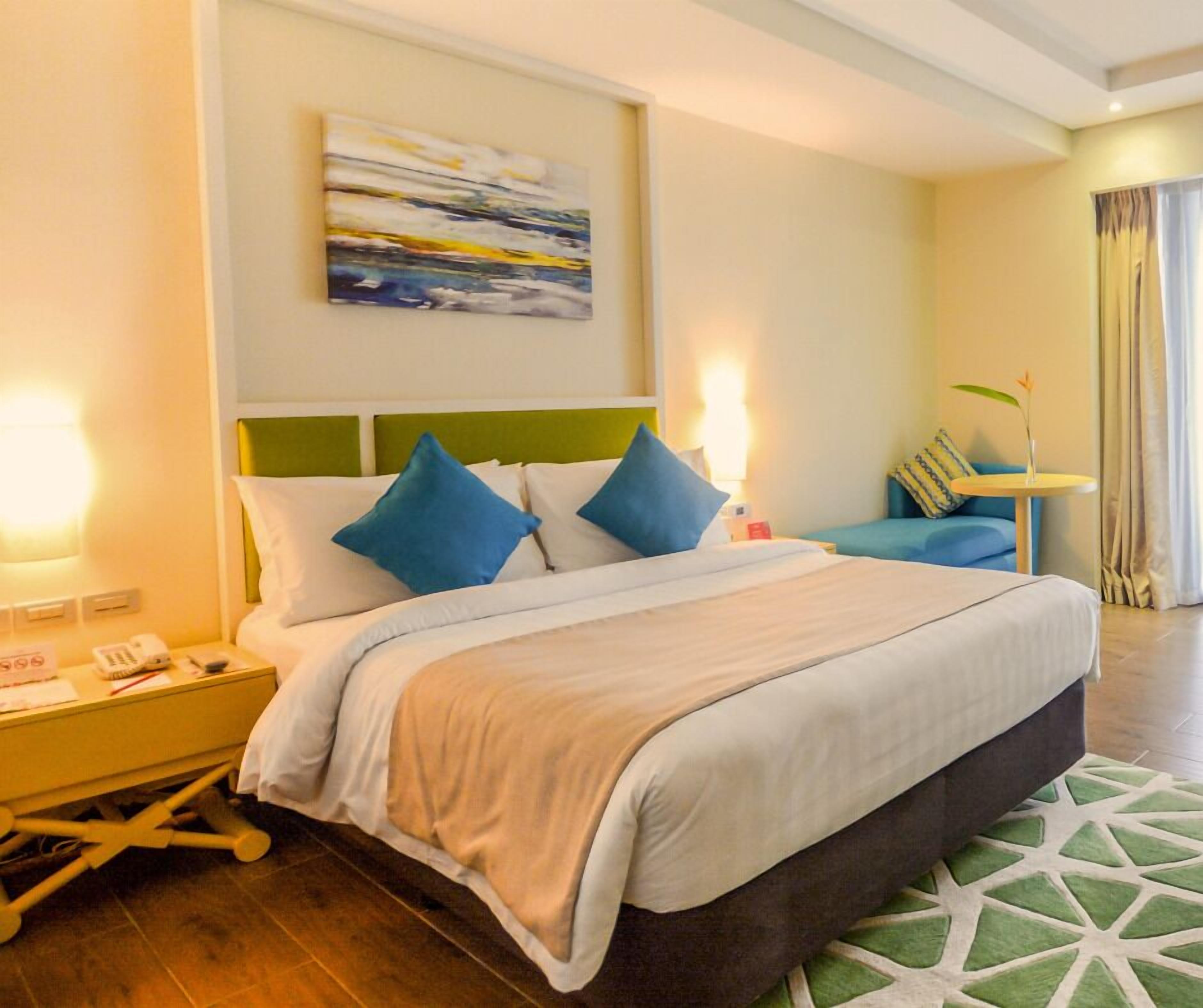 A hotel room with a bed, bedside tables, a small table with a vase, and a painting on the wall.