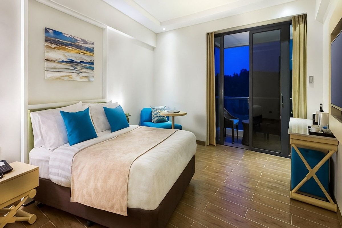 A hotel room with a bed, a desk, a chair, a TV, and a balcony with a view.