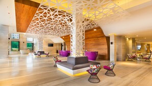 A spacious lobby with a modern design, featuring a central seating area with sofas and chairs, a wooden ceiling with geometric patterns, and large windows providing natural light.
