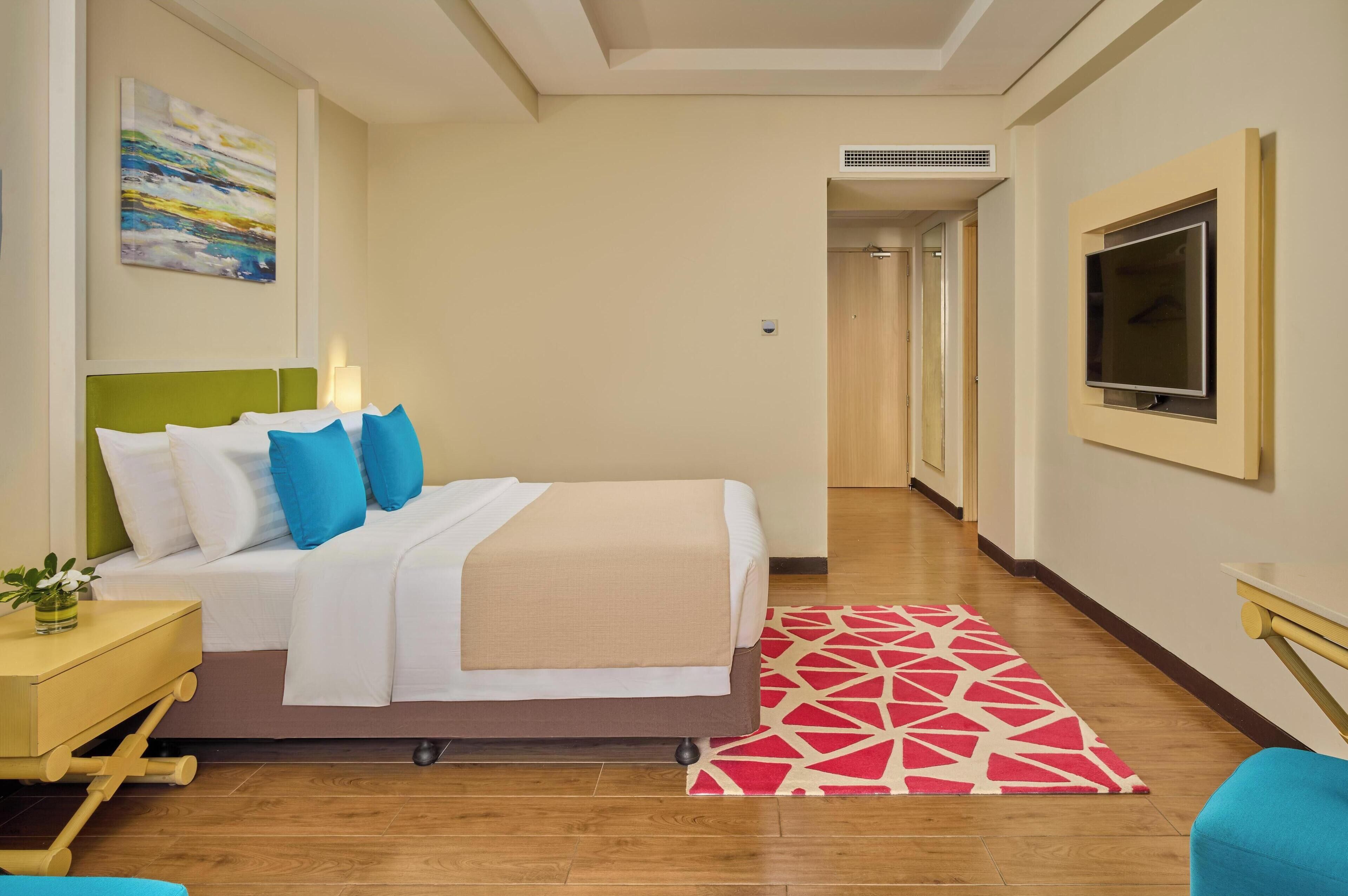 A hotel room with a bed, a TV, a painting, and a hallway.
