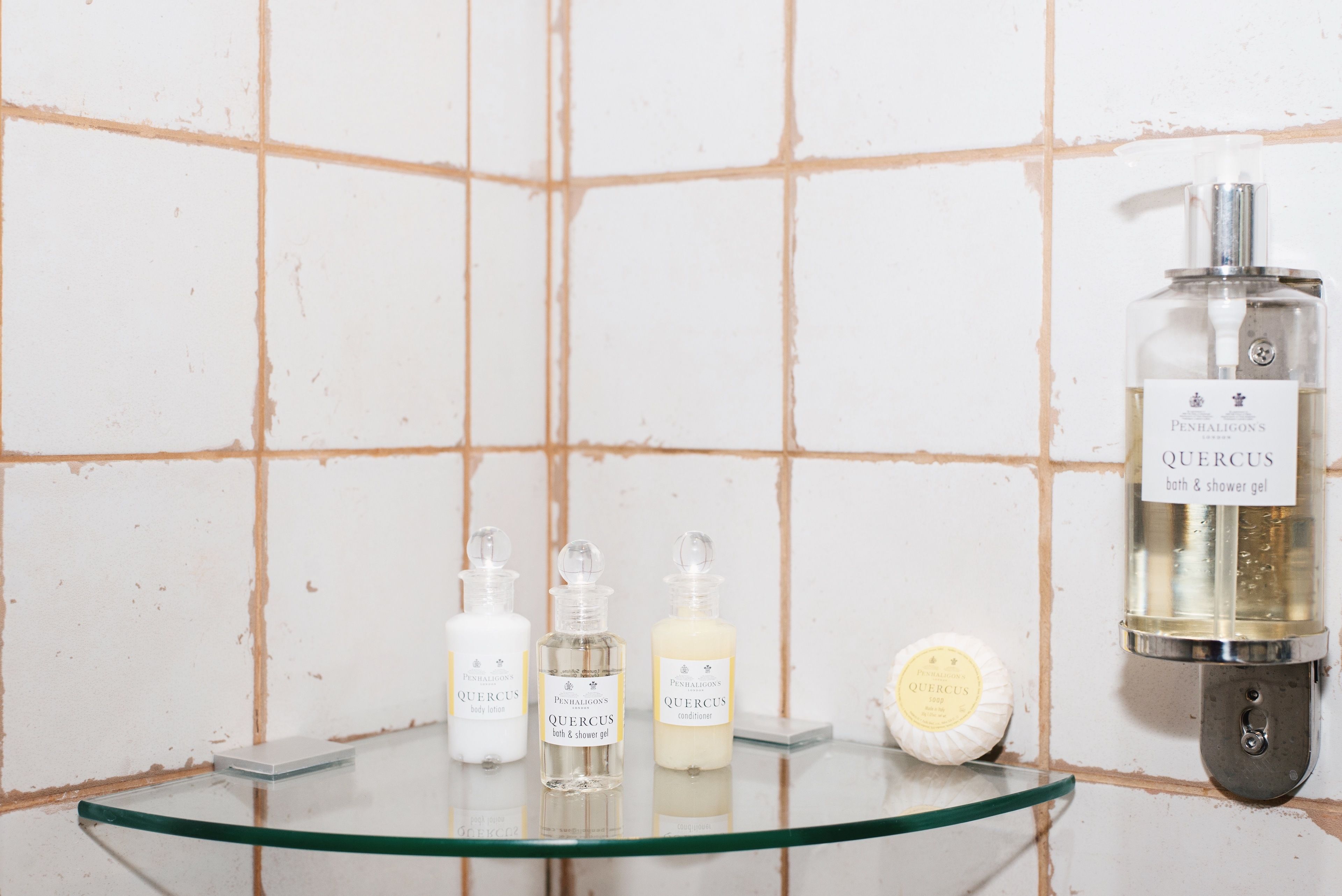 A bathroom with a glass shelf holding three bottles of Quercus body lotion, a Quercus bath & shower gel, and a Quercus cream bar.