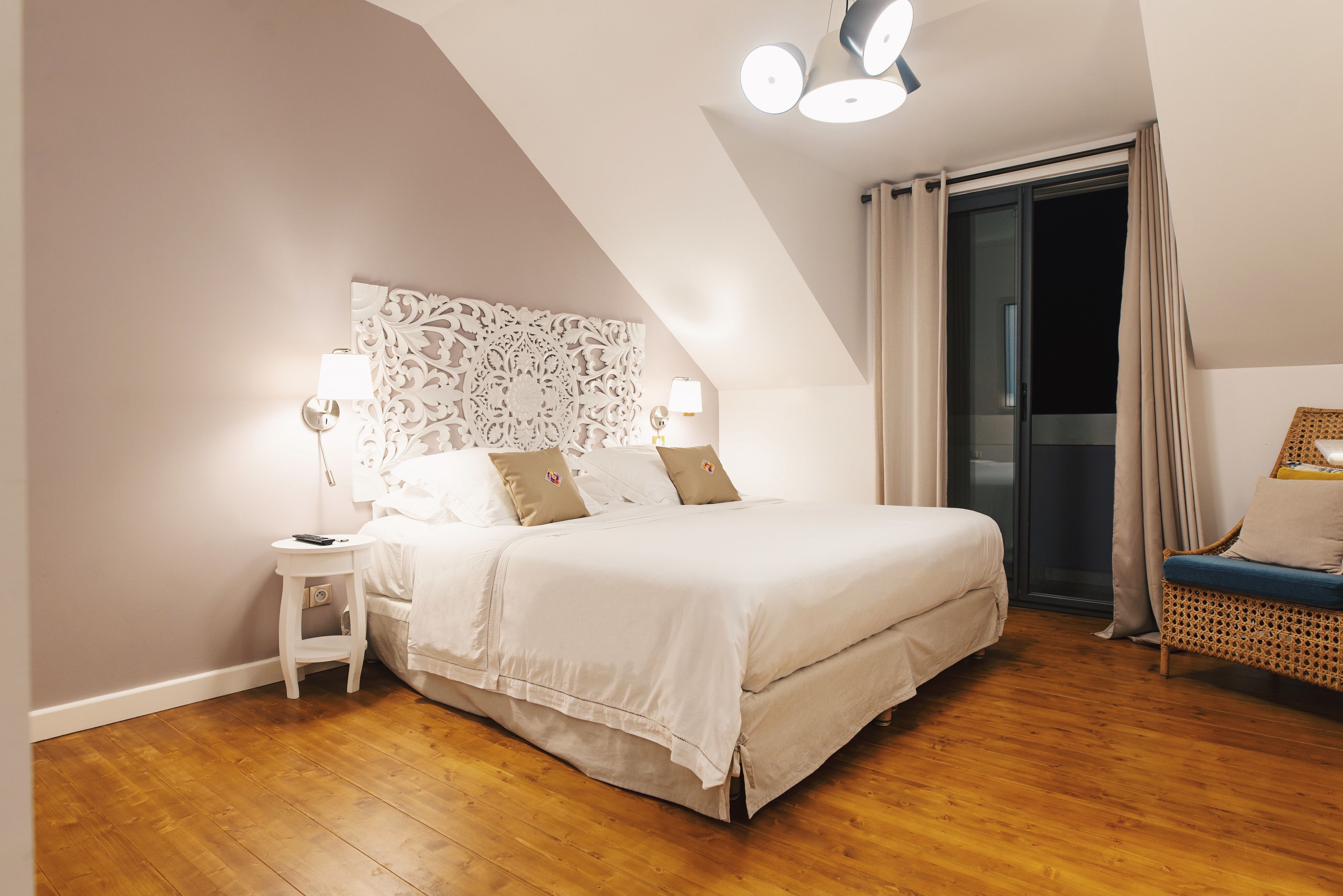 A bedroom with a bed, bedside tables, a decorative headboard, a wicker chair, and a sliding glass door.