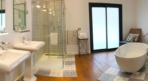 Un baño moderno con dos lavamanos, un box de ducha de vidrio, una bañera grande y una silla de mimbre junto a la ventana.