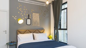 A neatly made bed with a blue bedspread and white pillows, a wall-mounted light fixture, and a decorative wall hanging. - Mr. Brooklyn - By TLV2GO (Tel Aviv)