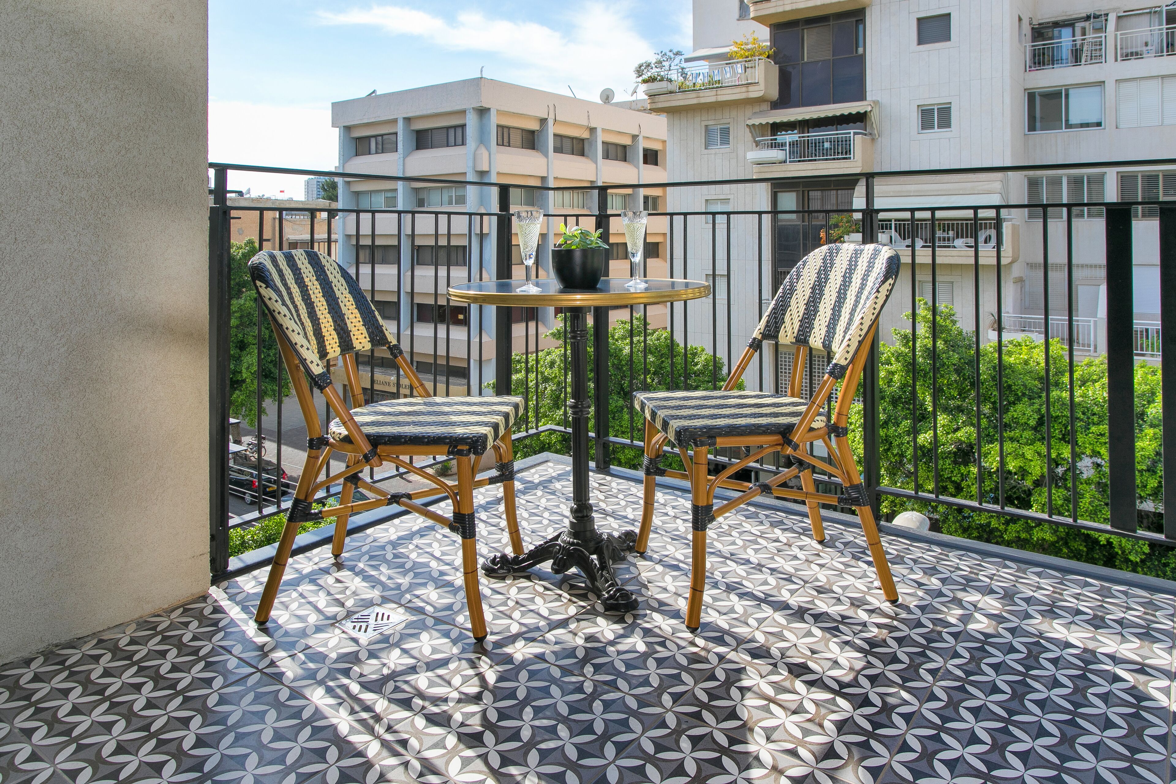 A balcony with two wicker chairs and a small round table, featuring a patterned floor and a view of surrounding buildings.