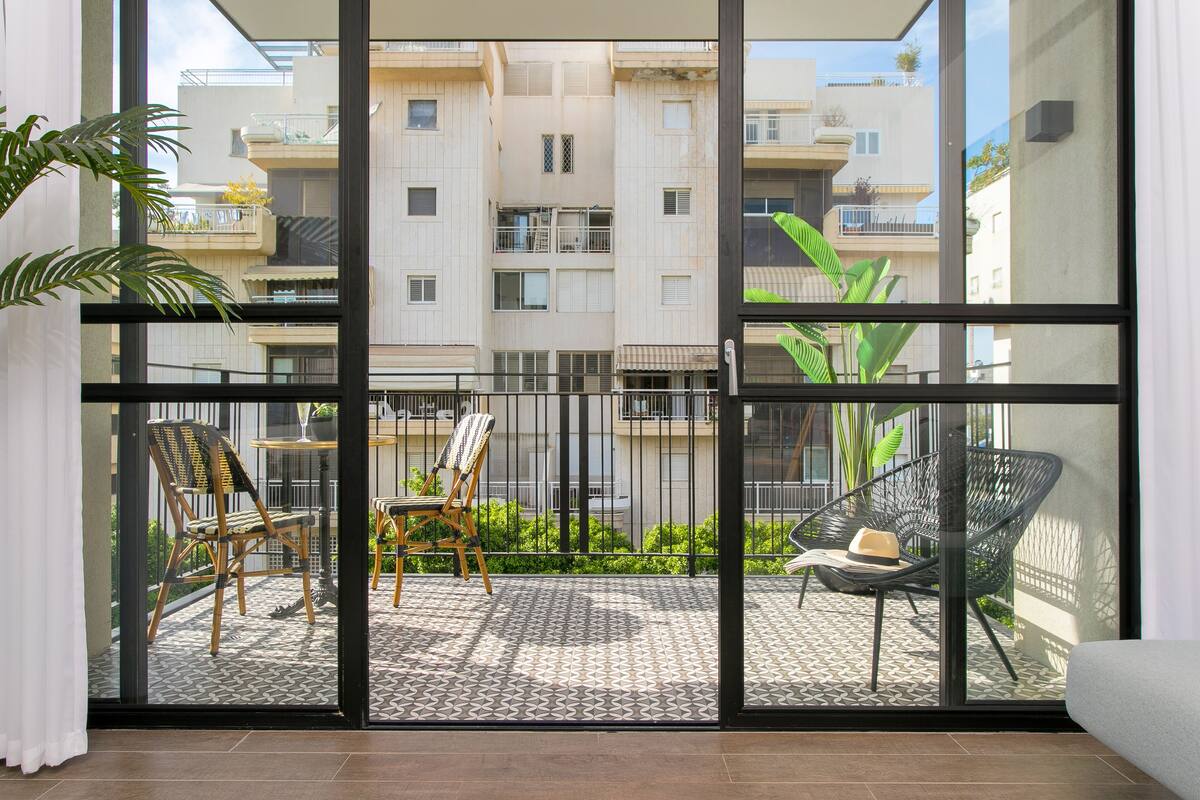 A balcony with two wicker chairs and a table, a patterned floor, and a view of a multi-story building with balconies.