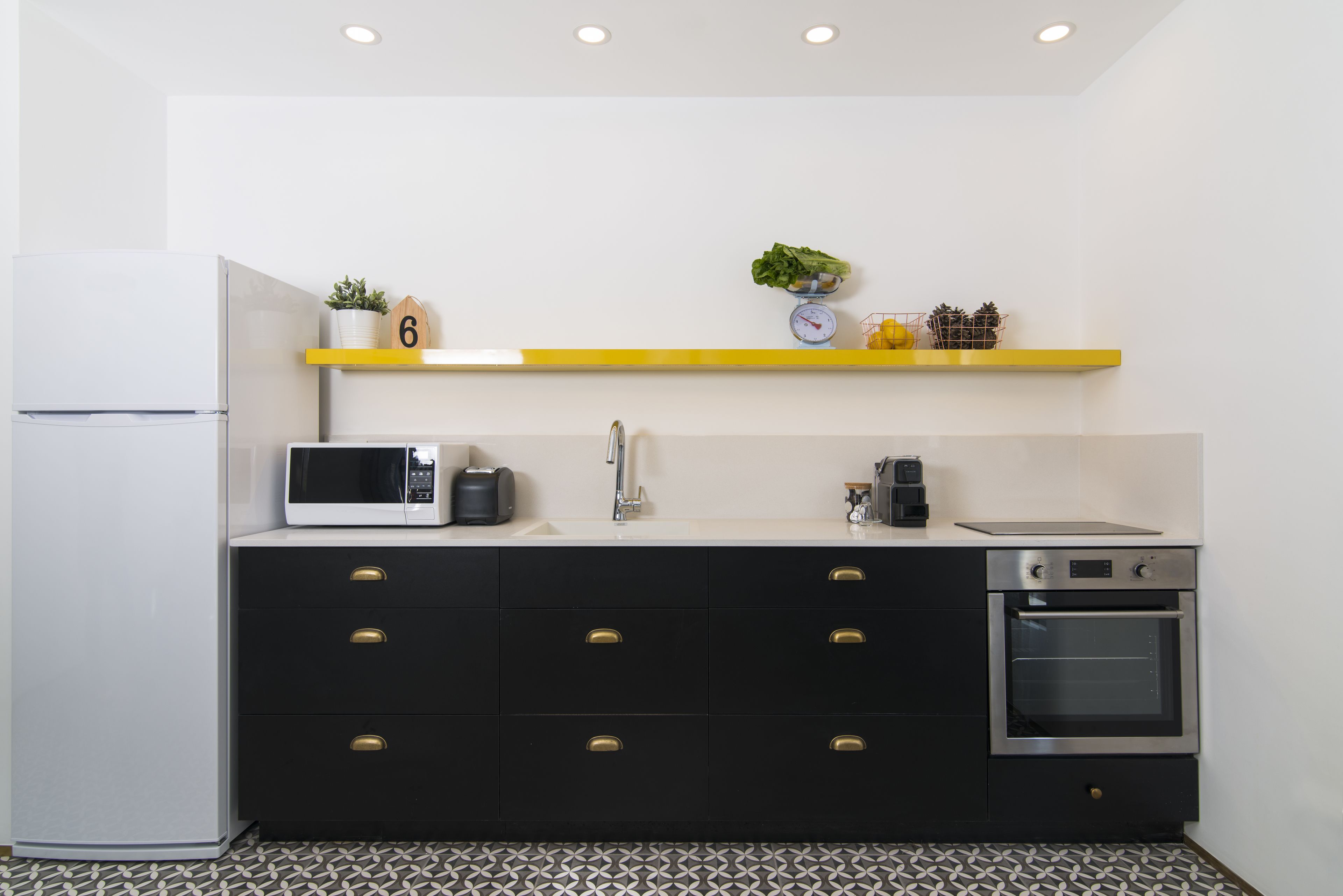 A compact kitchen with a microwave, stove, and oven. There is a sink with a faucet, a shelf with a scale and a plant, and a refrigerator.